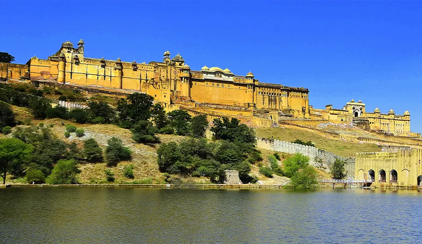 Large historic fort with yellow walls atop a hill, surrounded by greenery and overlooking a calm water body under a clear blue sky.