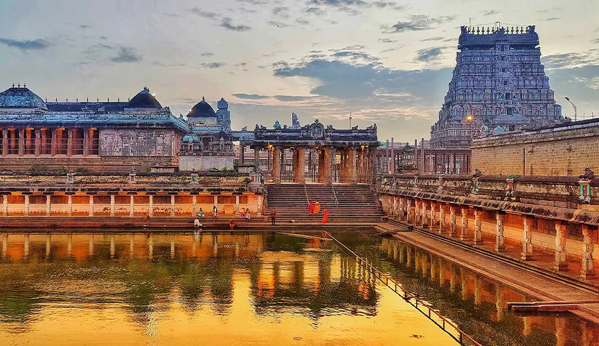 The image shows a famous South Indian temple complex with a large water tank, vibrant stone mandapam, and towering gopuram, all beautifully reflected in the golden-hued water at sunset.