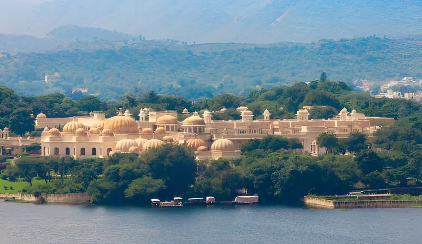 The image shows a grand palace with numerous domes and arches nestled among lush greenery, situated beside a serene lake with boats moored at the shore and hills in the background.