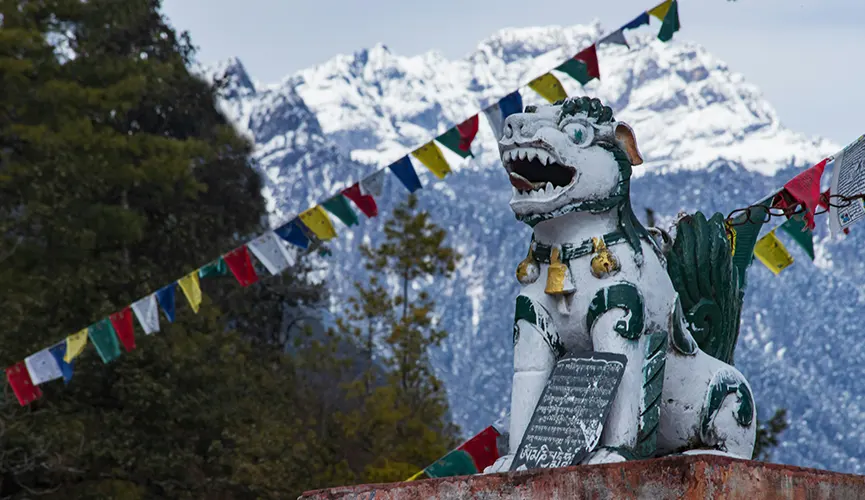 Statue of a guardian lion with colorful prayer flags, forest, and snow-capped mountains in the background.