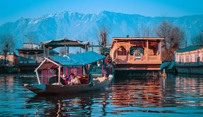 Traditional shikara boat with people gliding over a calm lake, wooden houseboats and snowy mountains in the background