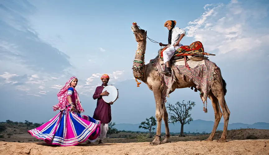 Man riding a decorated camel, with two people in vibrant traditional attire dancing and playing a drum in a desert landscape.