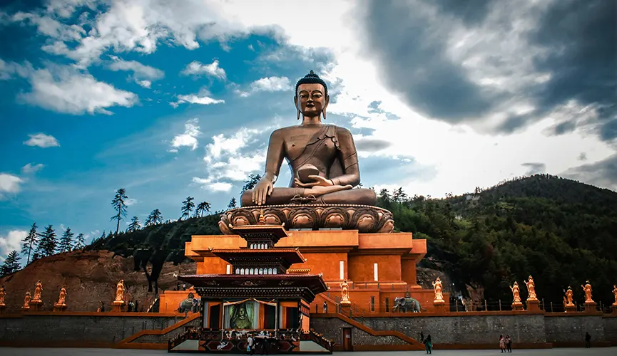 The image shows a massive seated Buddha statue atop a grand platform, set against dramatic clouds and forested hills. This is the Buddha Dordenma, a famous landmark in Thimphu, Bhutan, known for its serene presence and immense scale.