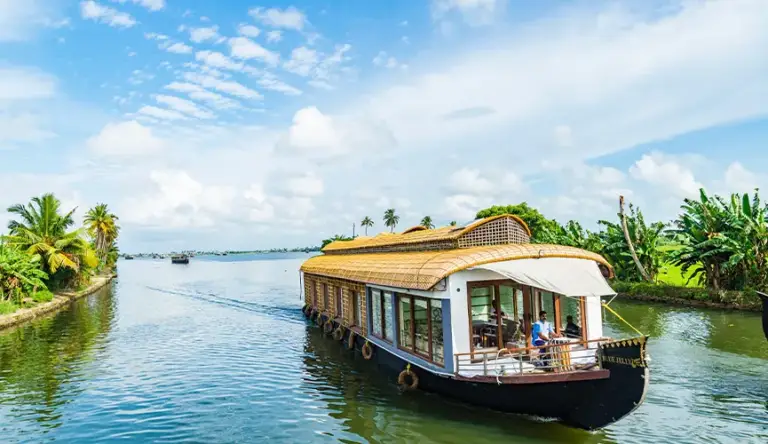 Scenic houseboat with people sailing on a lush, tropical river under a bright blue sky.