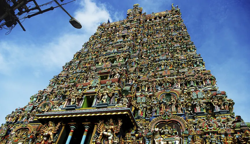 The image shows a towering, intricately decorated gopuram (gateway tower) of a South Indian Hindu temple, covered with hundreds of colorful statues of deities, mythical beings, and ornamental details against a bright blue sky.