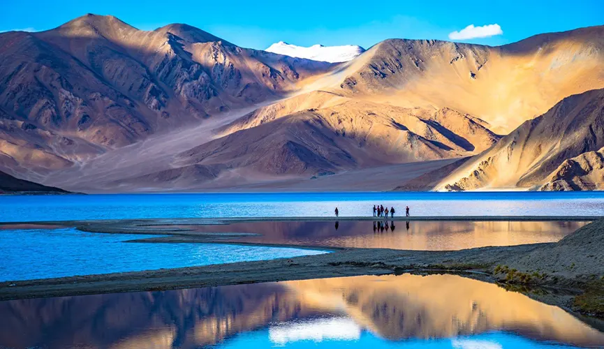Stunning blue lake surrounded by rugged mountains, with small figures standing on a strip of land reflected in the water.