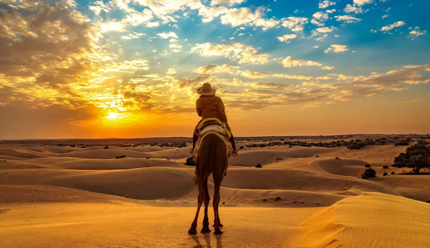 Person riding a camel across golden desert dunes at sunset under a vibrant sky.