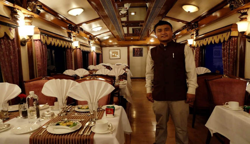 The image shows a formal dining car on a luxury train, set for a meal with white napkins elegantly fanned on the tables, fine china, and polished cutlery. A staff member in formal attire stands ready to assist passengers, contributing to the sophisticated and inviting atmosphere.