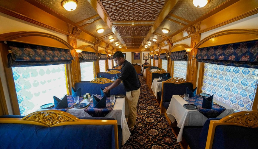 The image shows a luxurious dining car of a train, beautifully decorated with ornate seats, patterned carpets, and elegant table settings, as a staff member arranges napkins on the tables.