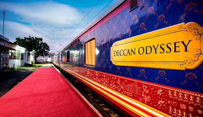 The image shows a lively traditional Indian welcome performance at a railway station, with dancers in colorful attire and a performer holding a large Ganesha puppet, set against the backdrop of an ornately painted luxury train.