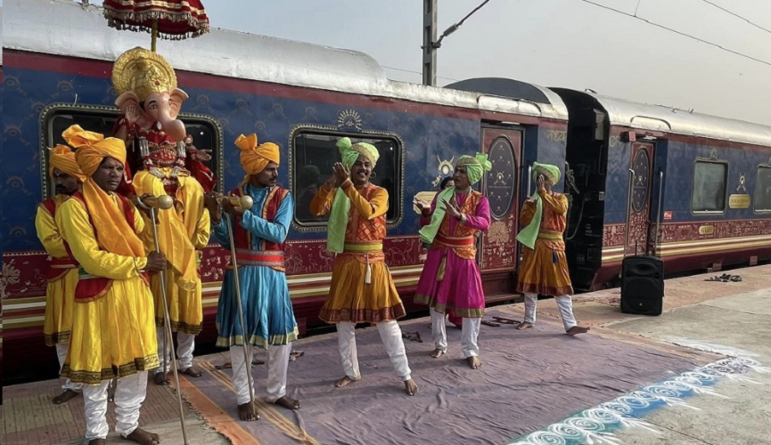 The image shows a lively traditional Indian welcome performance at a railway station, with dancers in colorful attire and a performer holding a large Ganesha puppet, set against the backdrop of an ornately painted luxury train.