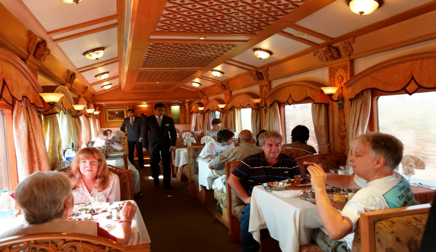 This image shows a beautifully decorated dining car of a luxury train where guests are dining at tables covered with white tablecloths. Ornate furnishings, elegant lighting, and attentive service create a refined atmosphere as passengers enjoy their meals while traveling.