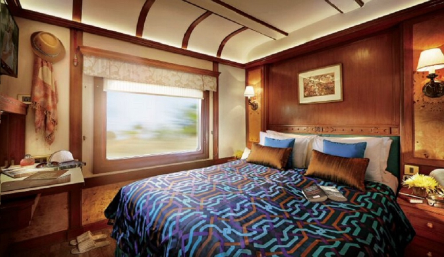 The image shows a luxurious train cabin with a large, comfortable bed, elegant wooden paneling, warm lighting, and a window revealing blurred scenery outside, suggesting the train is in motion.