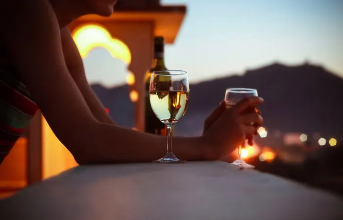 Close-up of a person holding two wine glasses on a balcony overlooking a city at sunset.