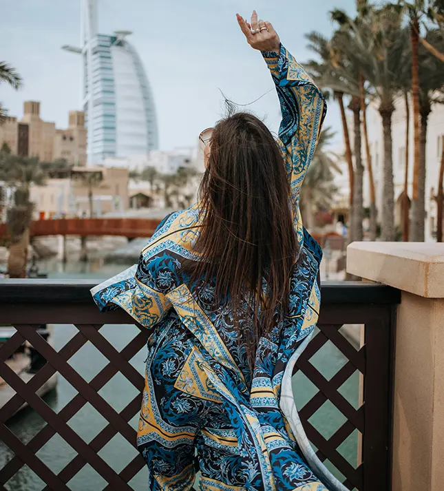 Woman in a blue patterned caftan posing with her arm raised, overlooking water with the Burj Al Arab hotel in the background.