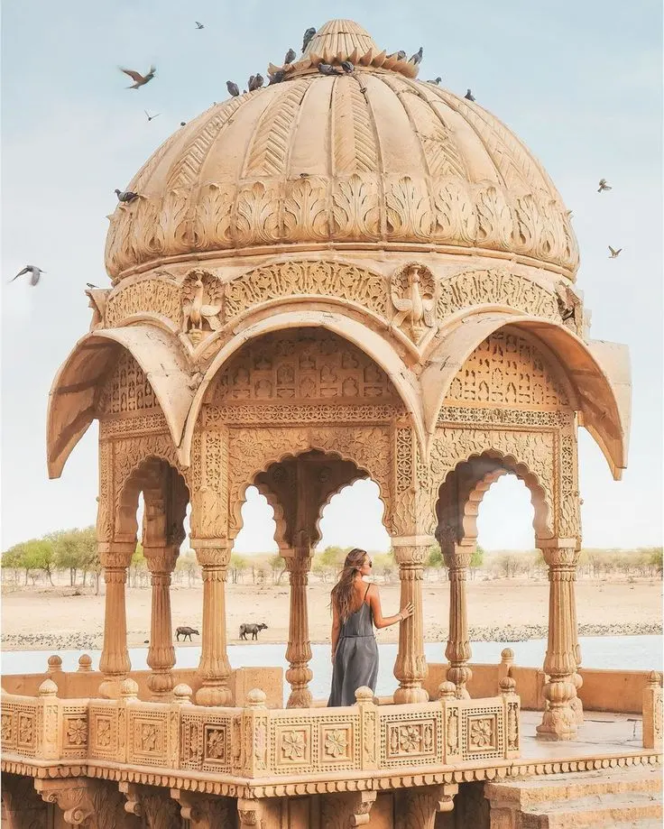 Woman standing inside a beautifully carved golden sandstone chhatri in Rajasthan, India, with pigeons flying overhead.