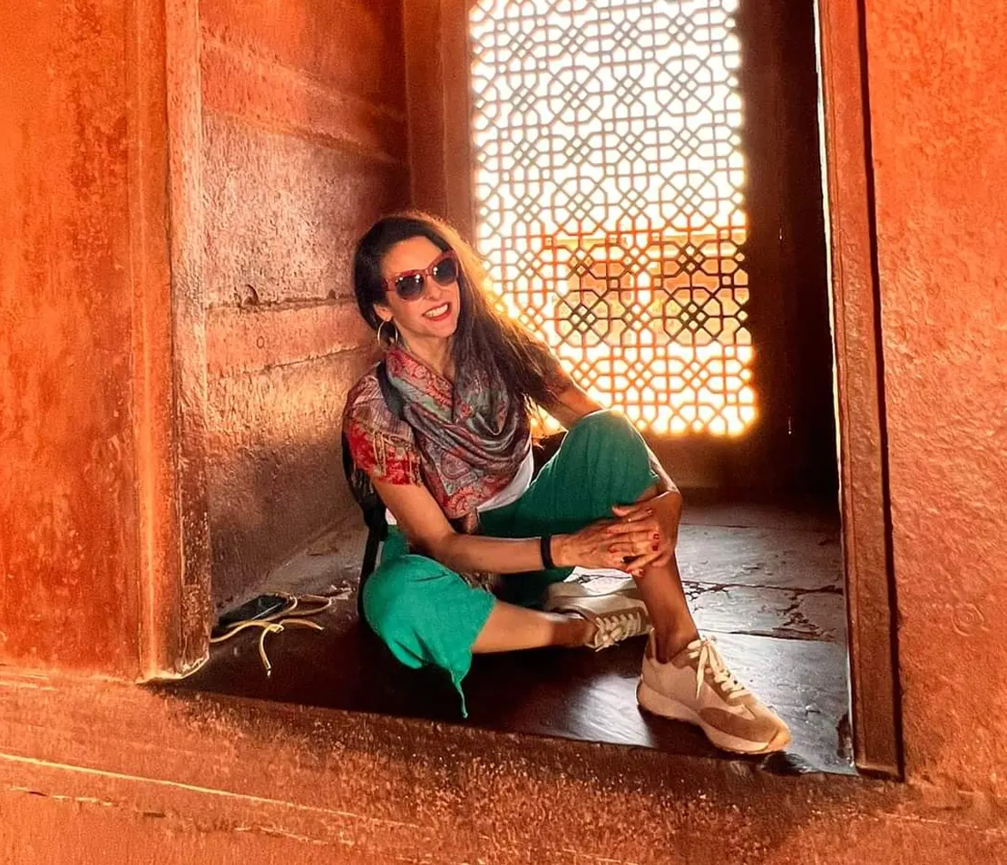 Smiling woman sitting in a red sandstone doorway with sunlight streaming through a jali screen.