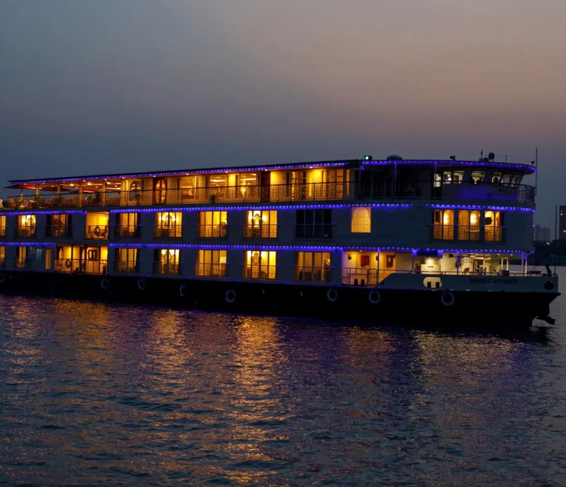 Large, multi-story river cruise ship illuminated with warm lights at dusk.
