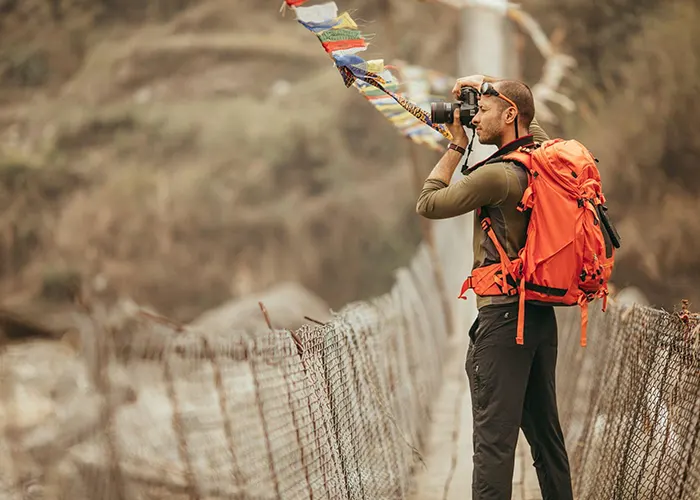 Hiker with an orange backpack taking a photo on a suspension bridge adorned with prayer flags.