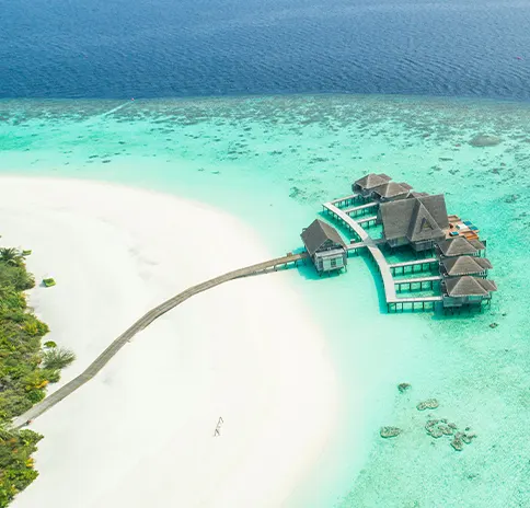 Aerial view of overwater bungalows connected by a jetty to a white-sand beach and turquoise lagoon in the Maldives.