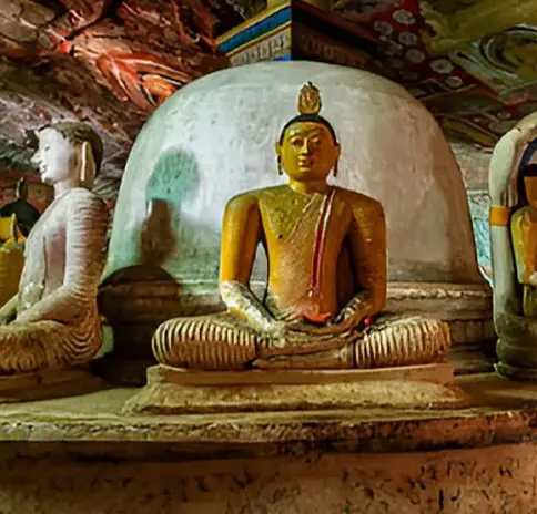 Seated Buddha statue with a yellow robe in front of a white dagoba inside a cave temple.