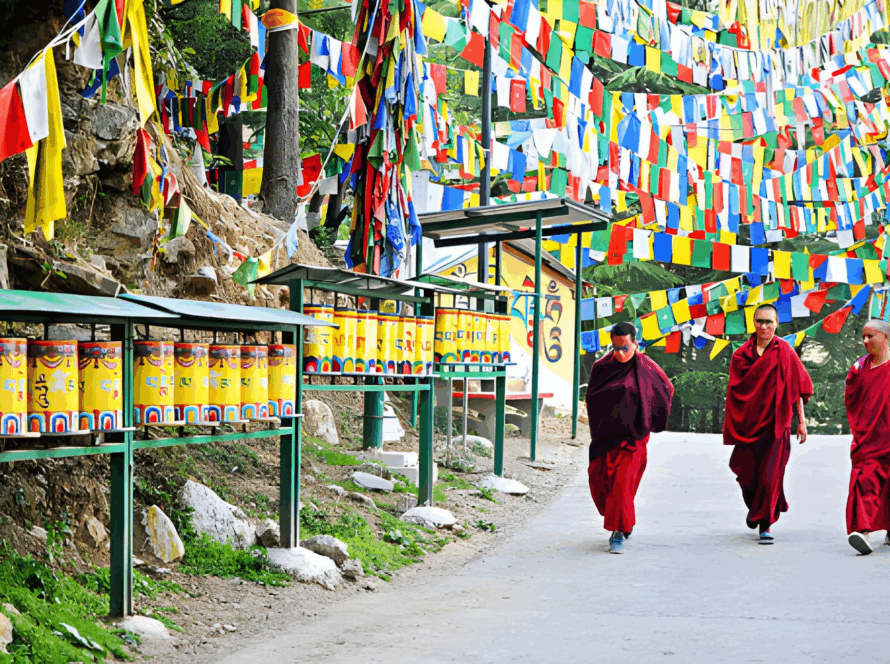  Buddhist monks in red robes walking past a row of yellow prayer wheels and numerous colorful prayer flags in McLeod Ganj, Himachal Pradesh.