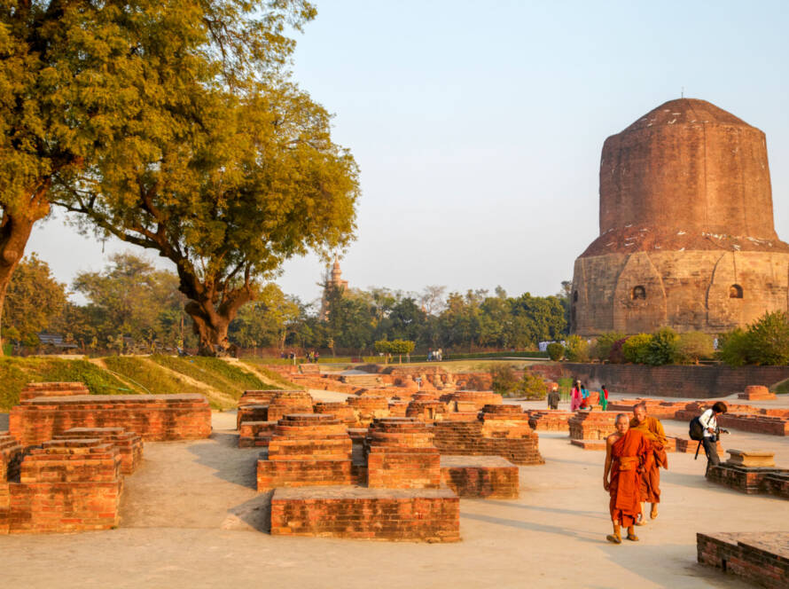 Sarnath: Buddhist monks walk among ancient brick ruins and Dhamek Stupa under large trees in historic pilgrimage site near Varanasi, India