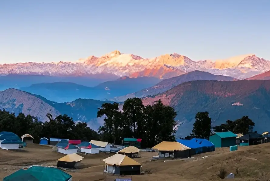 Panoramic view of a campsite in Chopta, Uttarakhand, nestled among hills with snow-capped Himalayan peaks glowing at sunrise, one of the Underrated Places in India.