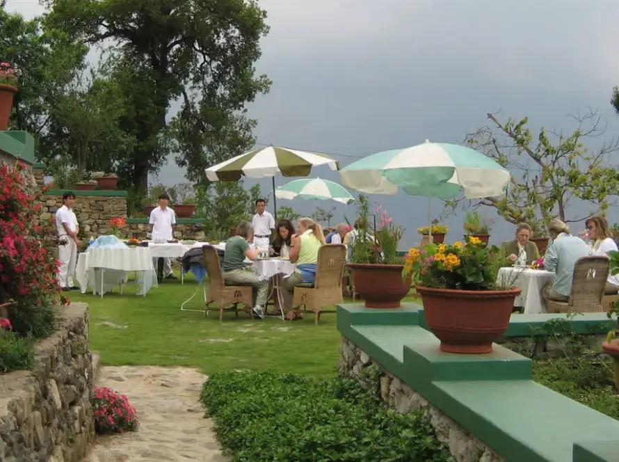 Outdoor garden restaurant on a cloudy day, showing patrons seated at tables under umbrellas and staff standing nearby, surrounded by stone walls and potted plants.
