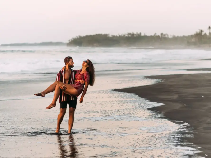 A man holding a woman in his arms while standing in the shallow water on a dark sand beach at sunset.