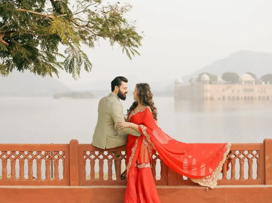 A couple in traditional Indian attire embracing on a red railing overlooking the Jal Mahal palace partially submerged in water in Jaipur.