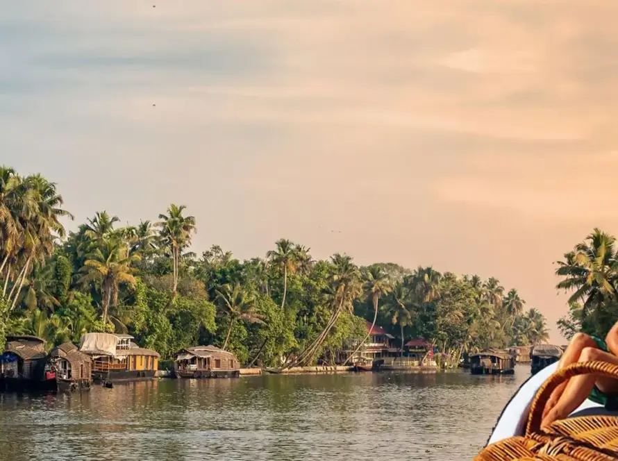 A couple sitting on the front of a boat, enjoying the view of houseboats and lush palm trees along the backwaters of Kerala at sunset.