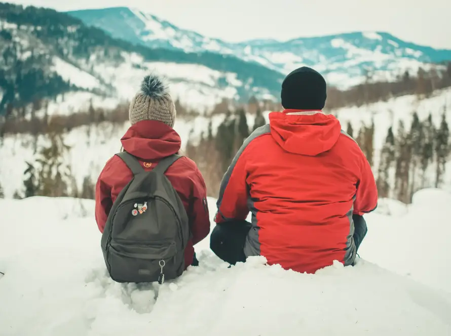 Two people in winter jackets sitting in the snow, looking out at a mountain range covered in snow and pine trees.