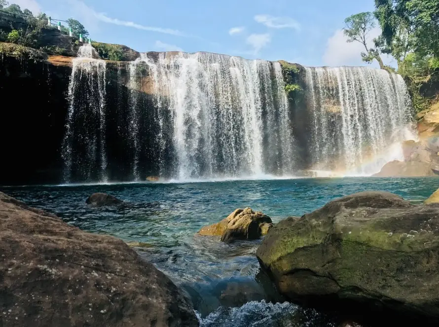 A wide waterfall cascading over a cliff face into a clear blue pool with large rocks in the foreground at Mawlynnong, Meghalaya, one of the Underrated Places in India.