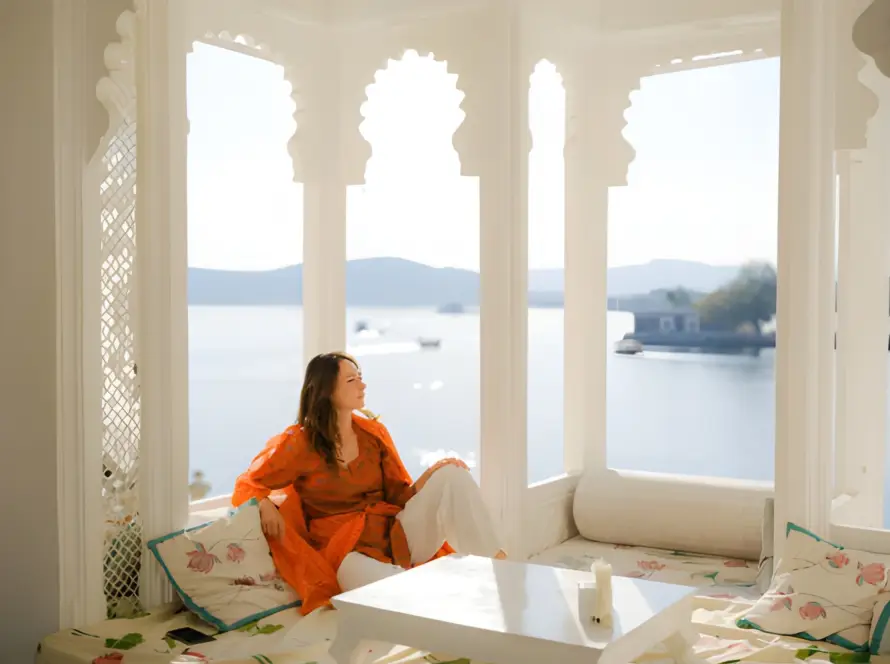 A woman in traditional attire relaxing in a bright, arched window seat overlooking a lake and distant palace in Udaipur, ideal for Honeymoon Destinations in India.