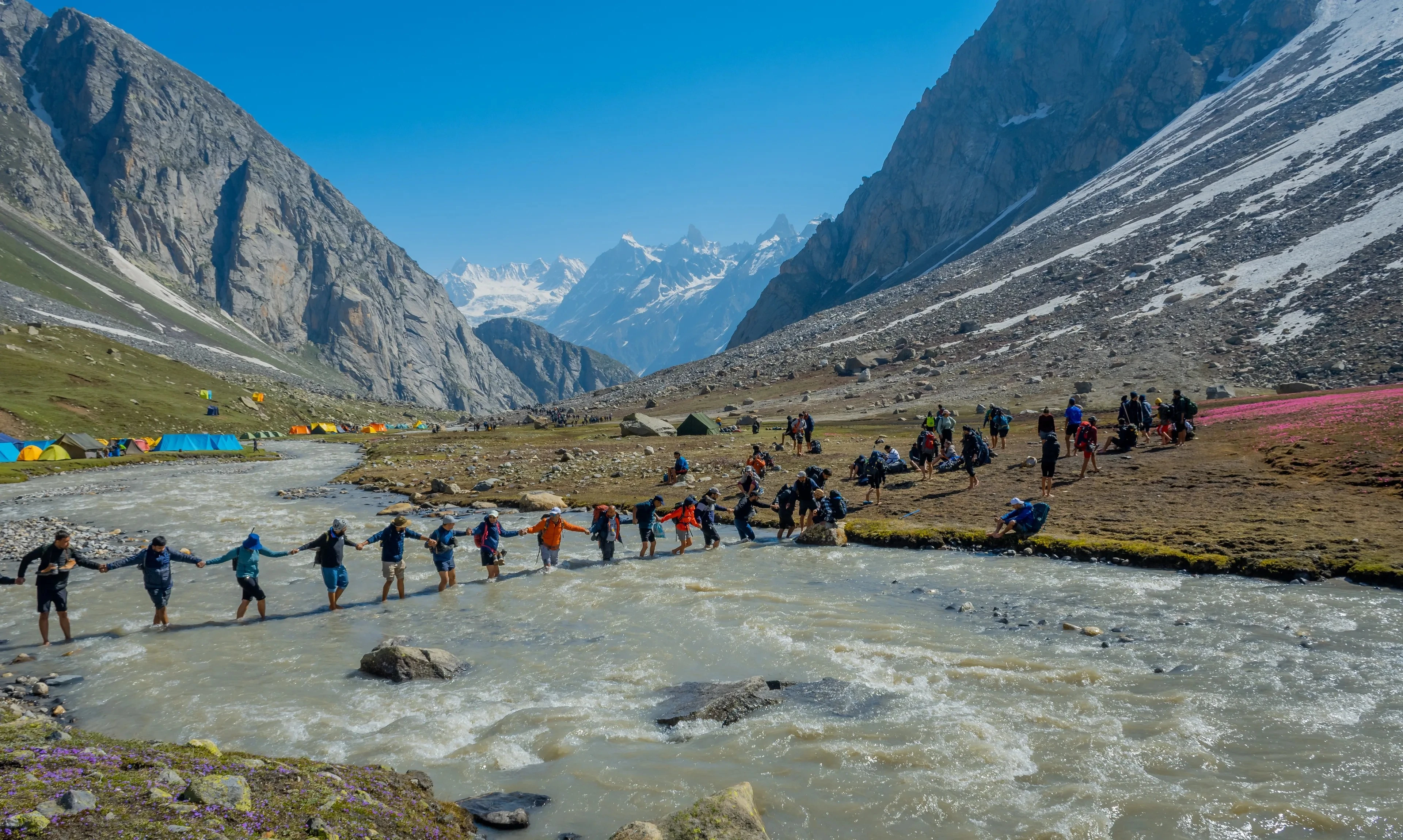 A long line of trekkers holding hands for balance as they wade across a shallow, fast-moving river in a rocky mountain valley, with colorful tents and jagged snow-capped peaks in the background.
