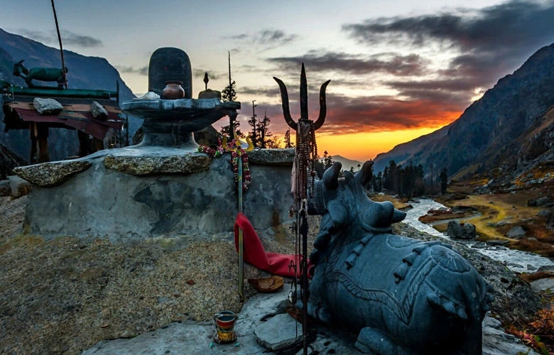 A religious shrine featuring a Shiva Lingam, Nandi statue, and trident overlooks a winding river valley during a vibrant sunset in the Har Ki Dun region.