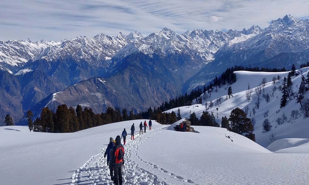 A line of trekkers follows a snow-covered ridge path towards a vast horizon of jagged, white peaks under a clear blue sky.