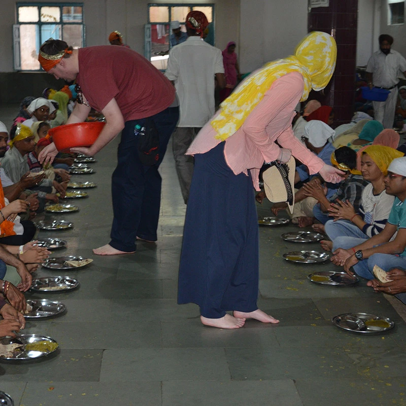 Langar seva volunteers serving free community meals at Gurudwara with people sitting on floor eating from metal plates 