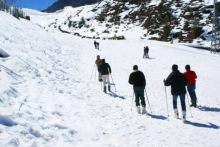A group of people wearing winter clothing and skis navigate a wide, snow-covered slope in a mountainous region.