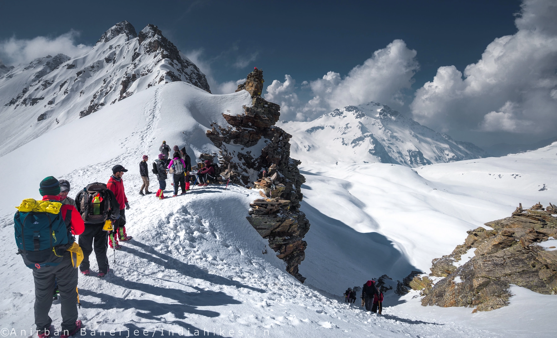 Several trekkers in winter gear sit and stand on a rugged, snow-covered mountain ridge under a deep blue sky. Jagged, white peaks and soft clouds are visible in the distance.
