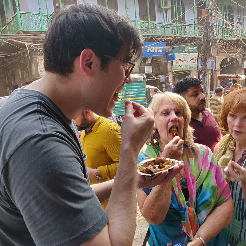 Old Delhi walking tour group tasting street food with local guide in narrow market alley with colorful buildings and shops 